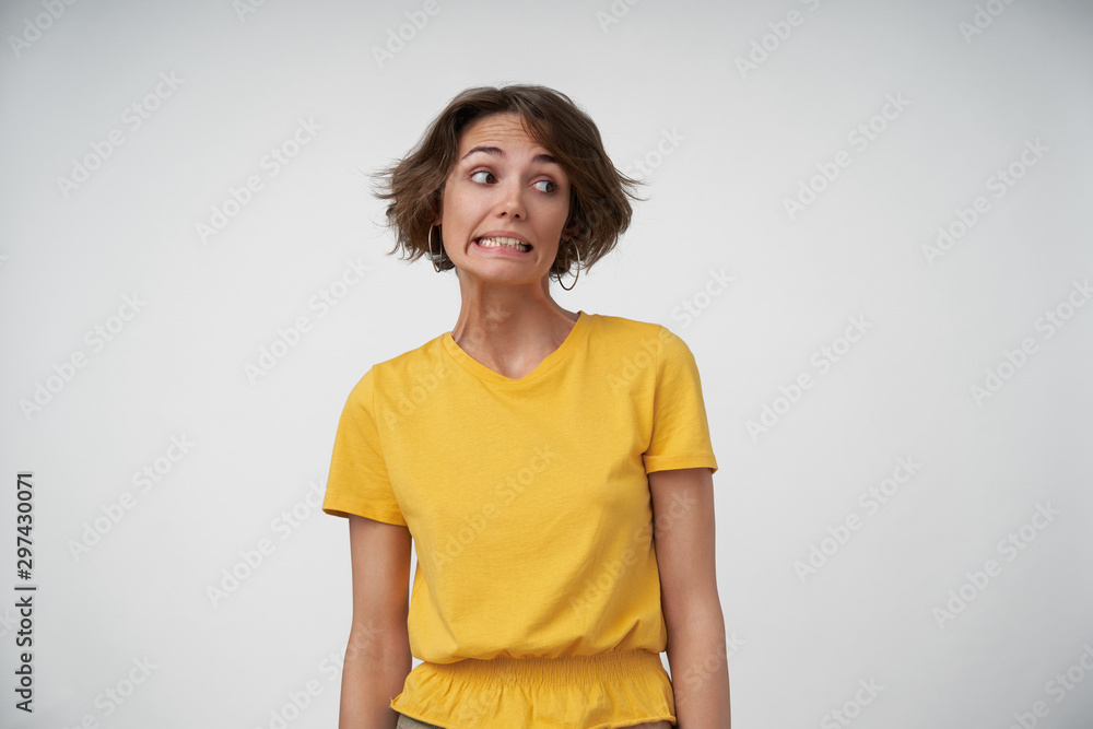 Portrait of embarrassed young female lady with short brown hair looking aside and showing teeth confusedly, keeping hands along her body while posing over white background Portrait of embarrassed young female lady with short brown hair looking aside and showing teeth confusedly, keeping hands along her body while posing over white background