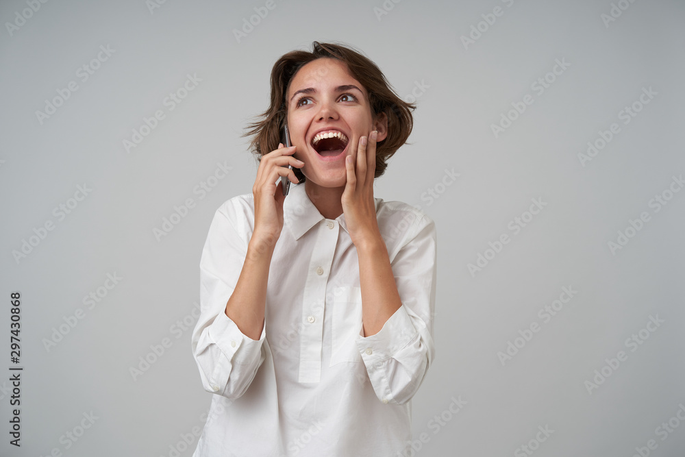Overjoyed attractive young woman with short brown hair talking on phone and receiving good news, raising happily her palm to face, standing over white background in formal clothes Overjoyed attractive young woman with short brown hair talking on phone and receiving good news, raising happily her palm to face, standing over white background in formal clothes