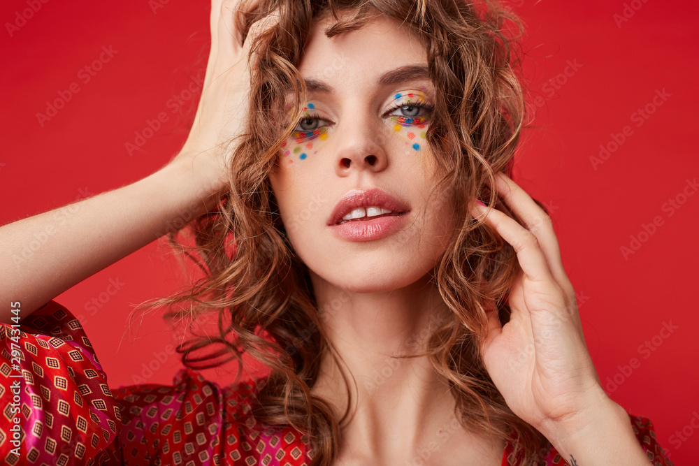 Attractive young curly woman with multicolored dots on her face looking at camera with calm face and touching her hair with raised hands, isolated over red background