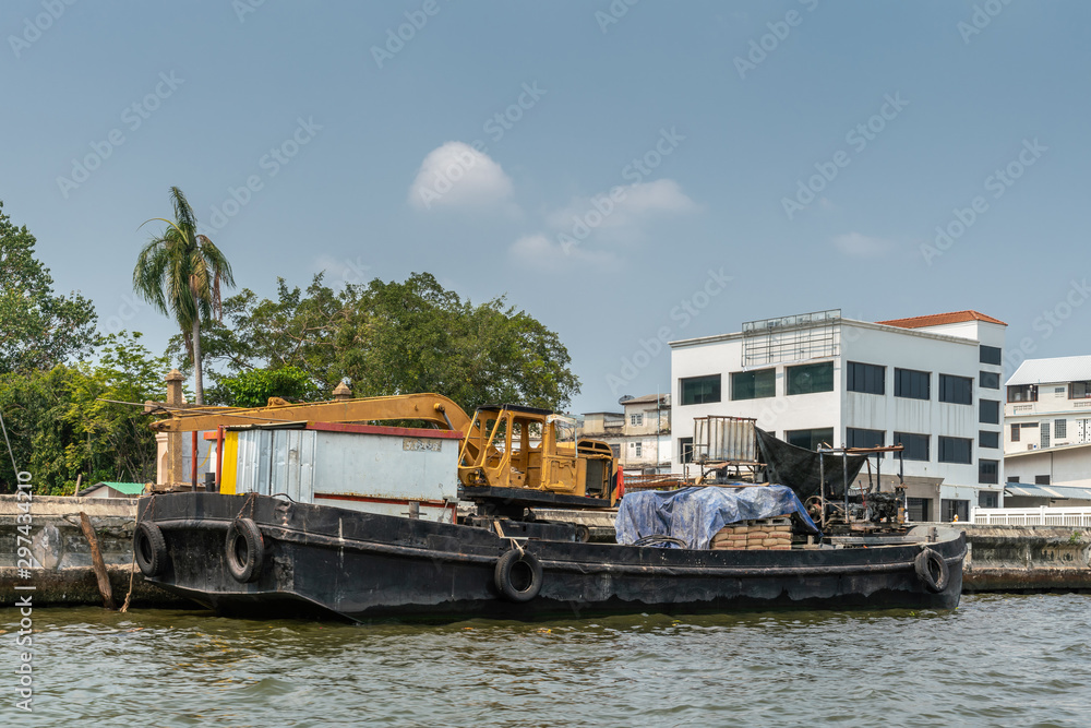 Bangkok city, Thailand - March 17, 2019: Bangkok Noi Canal. Small freighter barge with yellow crane and housing on top docked along the canal under blue sky. White building in back. 