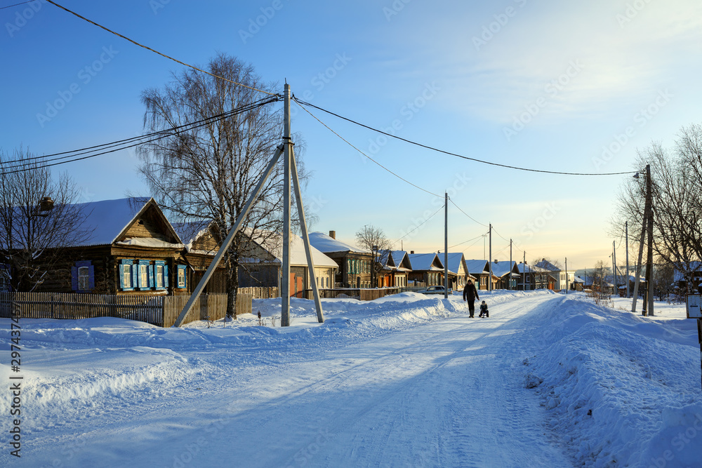 Russian village in winter. Man is carrying child on sled along snow-covered street. Village of Visim, Sverdlovsk region, Ural Mountains, Russia. Russian village in winter. Man is carrying child on sled along snow-covered street. Village of Visim, Sverdlovsk region, Ural Mountains, Russia.