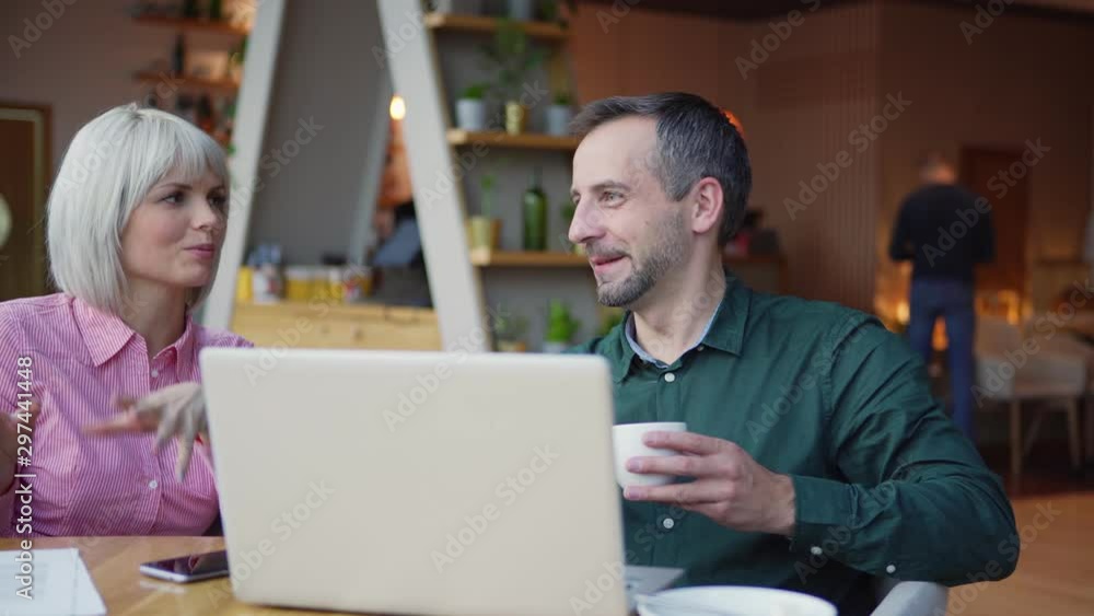 Camera panning left to adult man with cup of hot beverage speaking with female colleague in casual outfit while sitting at cafe table with laptop and working on project together Camera panning left to adult man with cup of hot beverage speaking with female colleague in casual outfit while sitting at cafe table with laptop and working on project together