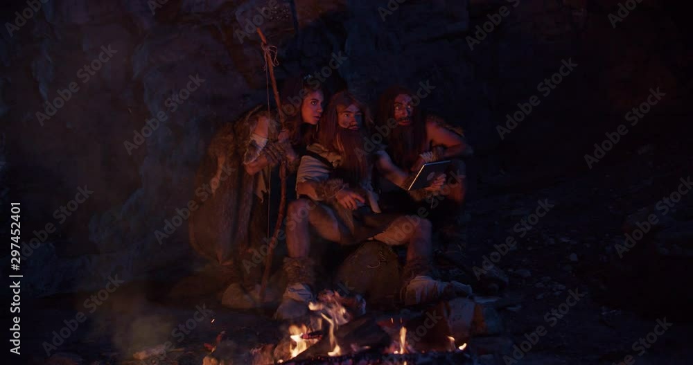 Close up cavemen hold bone with meat neanderthals wearing animal skin use tablet computer waving his hand having fun sitting by the fire in cave stone ancient evolution homo sapiens slow motion Close up cavemen hold bone with meat neanderthals wearing animal skin use tablet computer waving his hand having fun sitting by the fire in cave stone ancient evolution homo sapiens slow motion