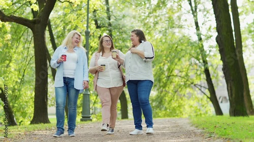 Full length shot of group of three middle aged plus size women walking down path in green park with takeaway coffee cups, talking joyfulle and laughing