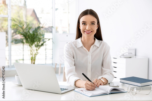 Young businesswoman writing in notebook while using laptop at table in office