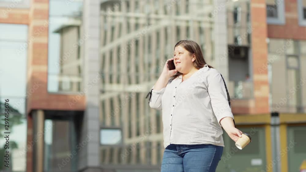 Tilt down three quarter length shot of overweight middle aged woman having unpleasant talk on cell phone and yelling during call, takeaway coffee cup in her hand Tilt down three quarter length shot of overweight middle aged woman having unpleasant talk on cell phone and yelling during call, takeaway coffee cup in her hand