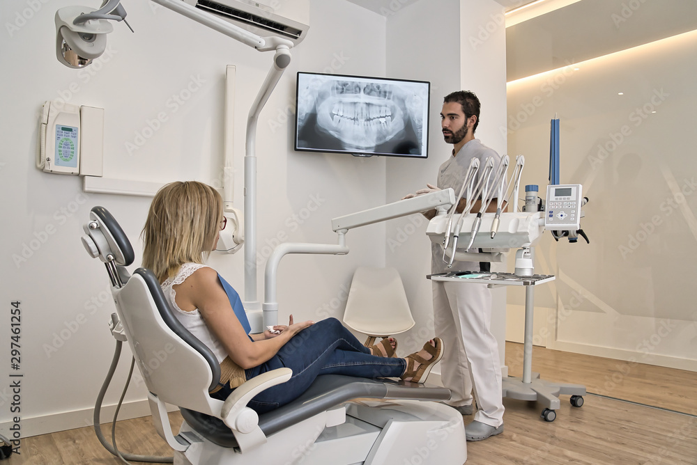 Black hair and beard dentist shows the client the x-ray of his mouth. Western client with blond hair was tumbling in the clinic chair. Modern dental clinic xray concept. - obrazy, fototapety, plakaty 