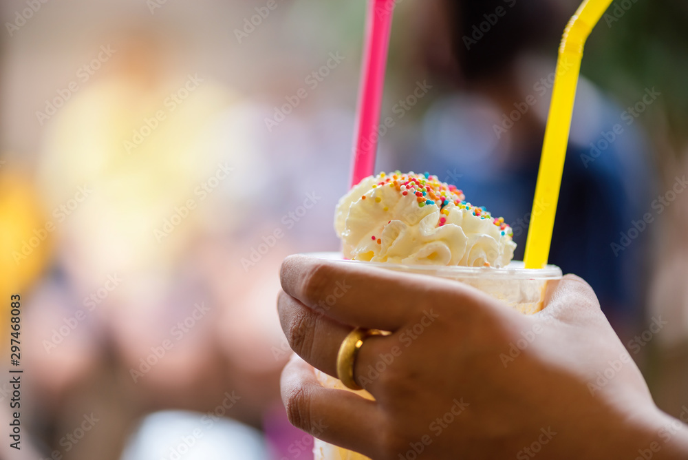 ็Hand holding cold sweet sprinkled with cream and candy flakes in plastic glass.