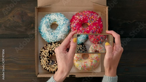 Top view - Woman's hands take pictures with smartphone of delicious fresh donuts in box on wooden table. 4k. Concept of food photo blogging. photo