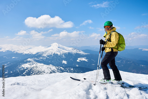 Sportsman skier in helmet and goggles with backpack standing on skis holding ski poles in deep white snow on copy space background of bright blue sky and highland landscape. Winter skiing concept.