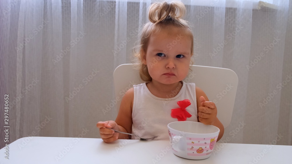 Infant girl eating baby food at a small childrens table on a white background. Nutrition and feeding of children. Infant girl eating baby food at a small childrens table on a white background. Nutrition and feeding of children.