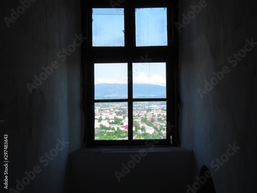 A window from a dark gloomy room into a bright sunny world. Old shabby window with dirty glasses  the dark walls and beautiful cityscape against the backdrop of the mountains outside