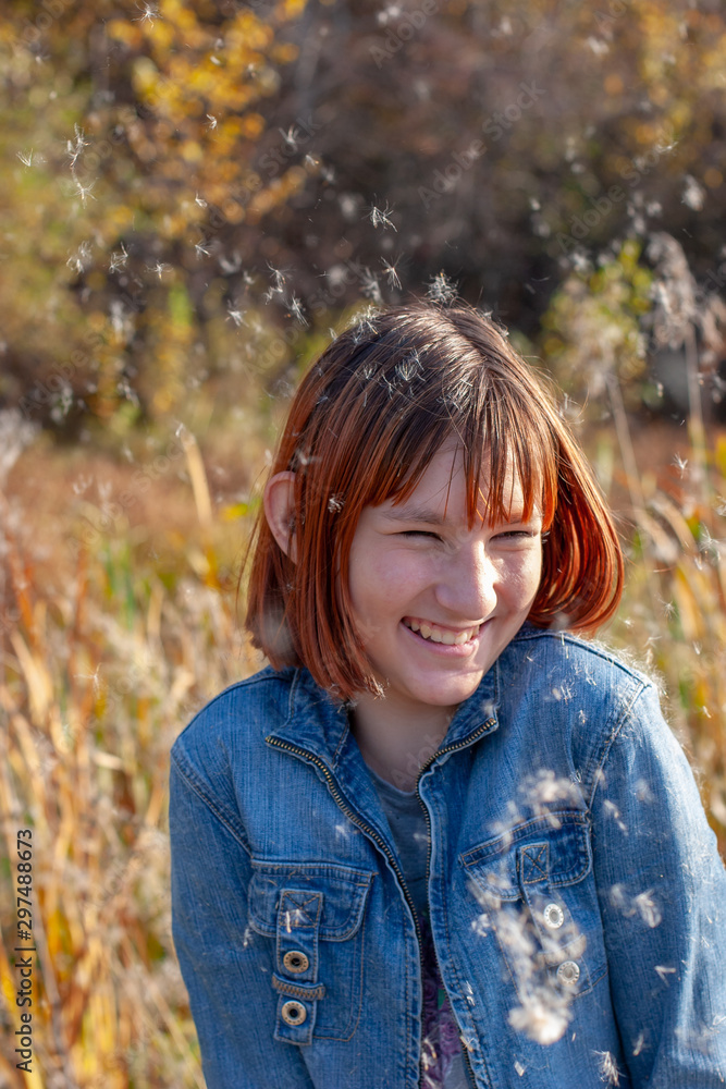A cheerful contented girl laughs and fluff from a cattail flies around. Selective focus on the girl. Autumn forest and bulrushes on a blurred background. Vertical. A cheerful contented girl laughs and fluff from a cattail flies around. Selective focus on the girl. Autumn forest and bulrushes on a blurred background. Vertical.