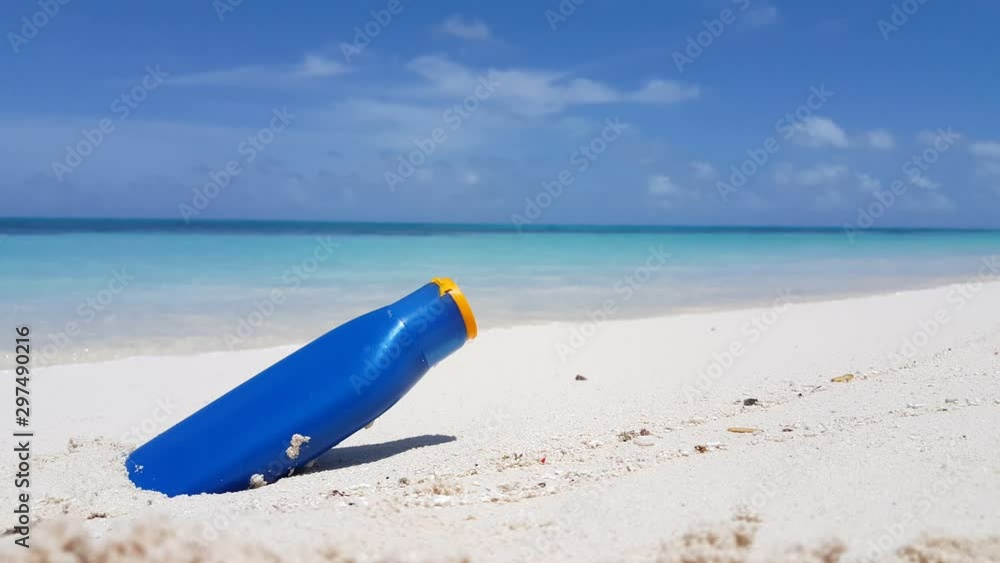 A sun screen bottle rests on white sand on a tropical beach with small waves washing onshore in Hawaii A sun screen bottle rests on white sand on a tropical beach with small waves washing onshore in Hawaii