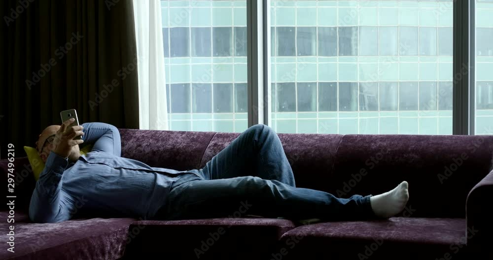 relaxed man is lying on couch in his home in daytime, swiping on display of smartphone relaxed man is lying on couch in his home in daytime, swiping on display of smartphone