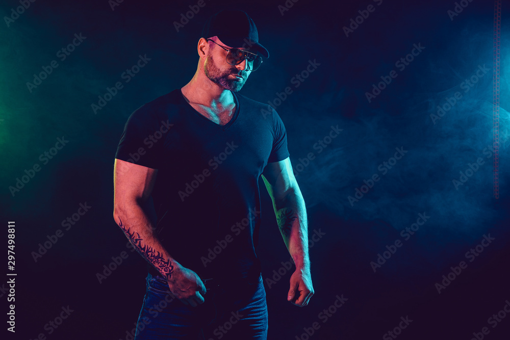 Naklejka premium Young strong man bodybuilder in cap on wall background. Dark dramatic colors. Smoke on background.