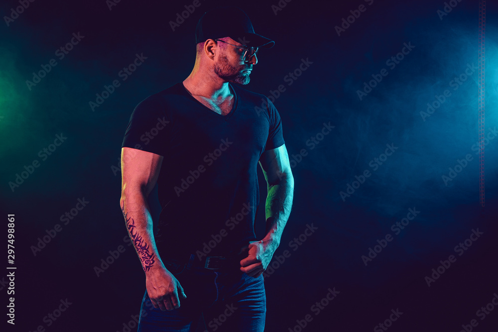 Naklejka premium Young strong man bodybuilder in cap on wall background. Dark dramatic colors. Smoke on background.