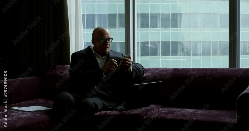 bald man is wearing business suit is sitting on couch and typing sms on cell phone bald man is wearing business suit is sitting on couch and typing sms on cell phone