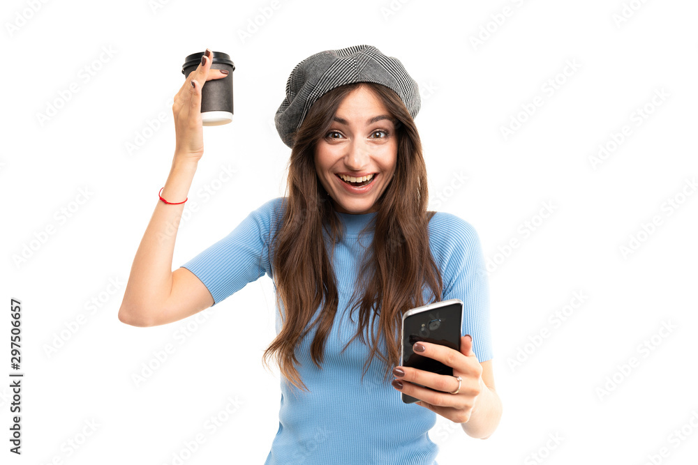 Young girl with delightful smile, long wavy chestnut hair, beautiful makeup, in blue jersey, black jeans, grey beret, with red bracelet stands with cup of coffee and phone in hand Young girl with delightful smile, long wavy chestnut hair, beautiful makeup, in blue jersey, black jeans, grey beret, with red bracelet stands with cup of coffee and phone in hand