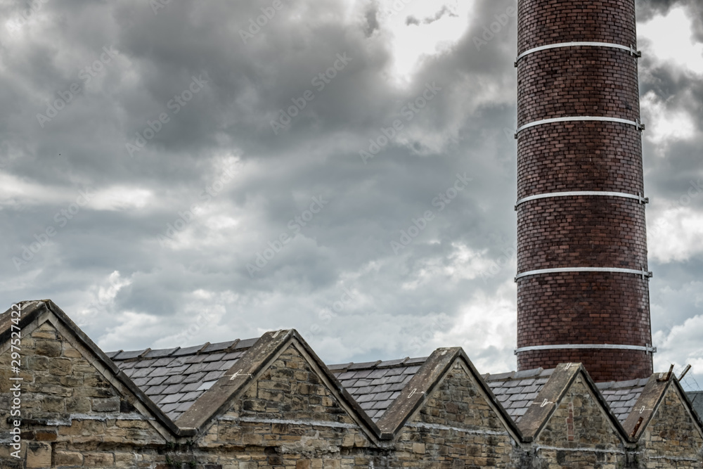 Old-style mill factory showing the individual roof structures. A large, brick built chimney is seen in the background, once used as a fire kiln. Old-style mill factory showing the individual roof structures. A large, brick built chimney is seen in the background, once used as a fire kiln.