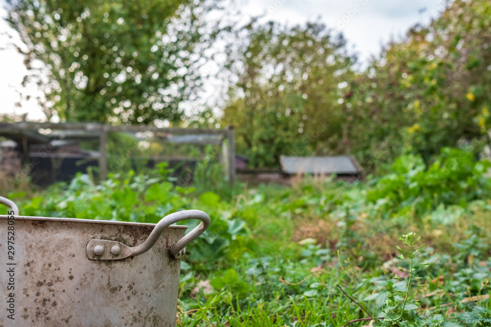 Shallow focus of a rural collection of timber chicken houses used for livestock, seen in an overgrown location. A nearby metal pot is a drinker pot. Shallow focus of a rural collection of timber chicken houses used for livestock, seen in an overgrown location. A nearby metal pot is a drinker pot.