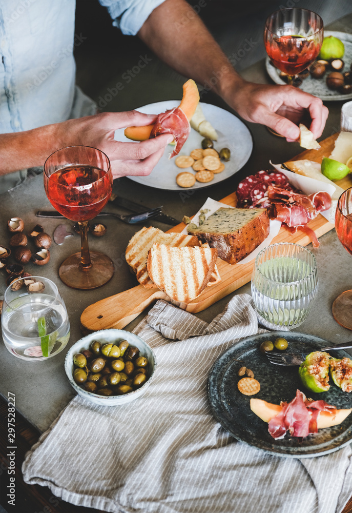Mid-summer picnic with wine and snacks. Charcuterie and cheese board, rose wine, nuts, olives, fresh fruits and mans hands with food over concrete table background, top view