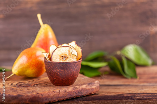 Homemade dried fruit chips in wooden cup, two fresh pears on wooden background with green leaves. Concept of healthy and vegan food. Copy space.