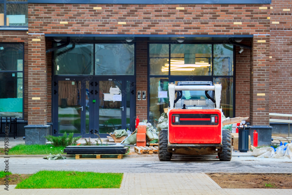 Fototapeta premium Skid loader stands in front of entrance to new multi-storey house and waits for garbage to be loaded into bucket. Mini bulldozer waiting for cargo in front of porch