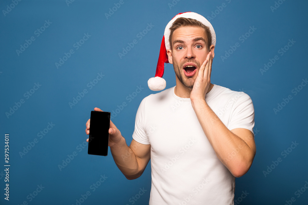 shocked young man in santa hat holding smartphone with blank screen isolated on blue shocked young man in santa hat holding smartphone with blank screen isolated on blue