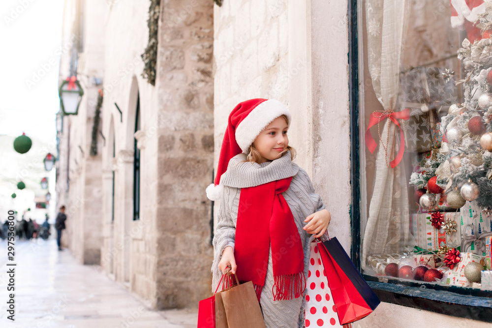 Christmas shopping. Child girl with paper bags on market street. Cute kid in red santa hat makes purchases by store showcase decorated with gifts, Christmas tree. Cozy fair, New Year and Black Friday