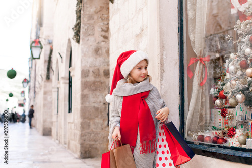 Christmas shopping. Child girl with paper bags on market street. Cute kid in red santa hat makes purchases by store showcase decorated with gifts, Christmas tree. Cozy fair, New Year and Black Friday © Marina April