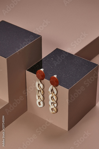 Object photo of an ivory plastic chain links earrings with brown round base, located on a dark beige cube with black marble upper side. The space around cube is polyformed. photo