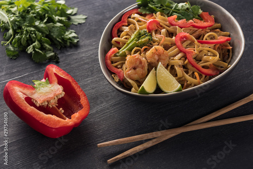 selective focus of tasty spicy seafood thai noodles near chopsticks, paprika and parsley on wooden grey surface