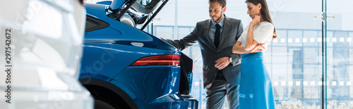 panoramic shot of attractive woman and handsome man looking at car trunk in car showroom