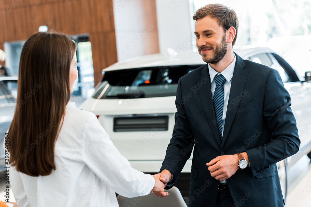 happy car dealer shaking hands with woman in car showroom