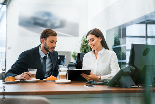 selective focus of happy car dealer holding clipboard near man and tasty croissants