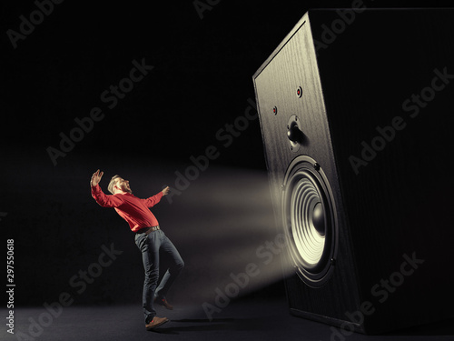 The sound wave from big sound speaker set back a screaming man in red shirt on black background. Image.
