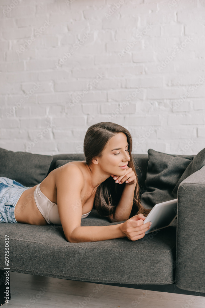 smiling woman in white bra and jeans using digital tablet on sofa in living room smiling woman in white bra and jeans using digital tablet on sofa in living room