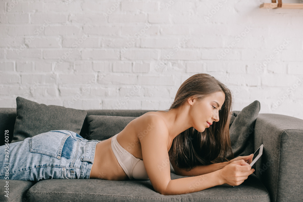 beautiful woman in white bra and jeans using digital tablet on sofa in living room beautiful woman in white bra and jeans using digital tablet on sofa in living room