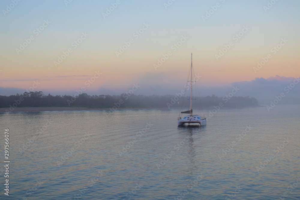 Big Catamaran Sail Boat Moored In A Calm Blue Harbour At Sunrise With Low Clouds In The Orange Sky