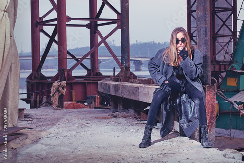 Obraz na płótnie Armed girl in a trenchcoat lights up a cigarette while waiting at a rendezvous point under the bridge near a river
