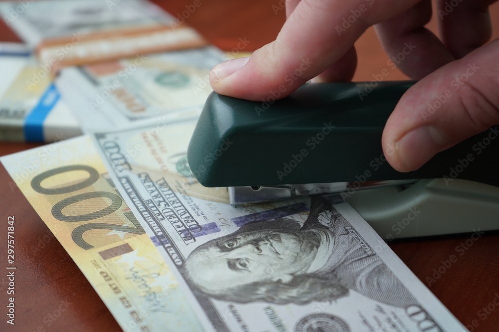The stapler fastens the dollar and euro notes with iron clips. Stapler with paper clips on the background of banknotes. The stapler fastens the dollar and euro notes with iron clips. Stapler with paper clips on the background of banknotes.