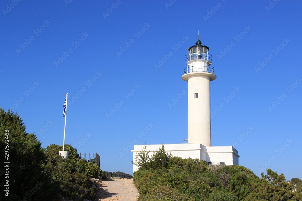 Beacon of Lefkada island south cape stone built lighthouse tower at summer, clear blue sky at background. Pathway and Greek flag at foreground Beacon of Lefkada island south cape stone built lighthouse tower at summer, clear blue sky at background. Pathway and Greek flag at foreground