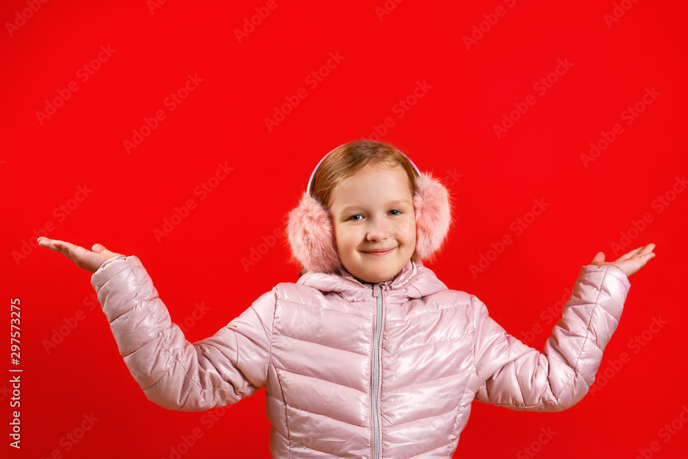 The little girl raised her hands up. A child in a jacket and warm ear muffs on a red background. The concept of happiness, winter The little girl raised her hands up. A child in a jacket and warm ear muffs on a red background. The concept of happiness, winter