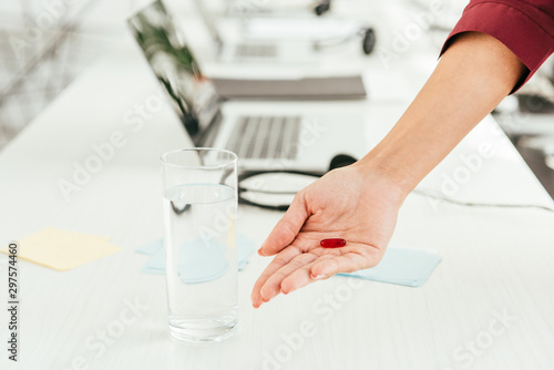cropped view of broker holding pill near glass of water and laptop in office