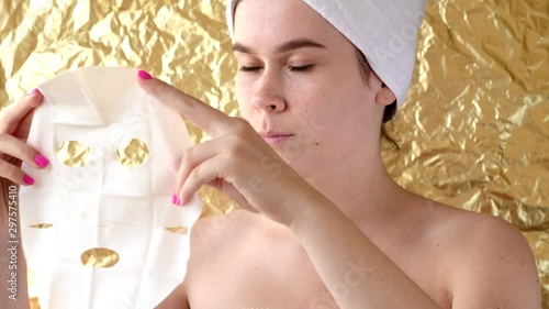 Young woman apply cosmetic fabric facial mask on golden background. Face moisturizing mask with charcoal, spa beauty treatment, skincare, cosmetology. Close up