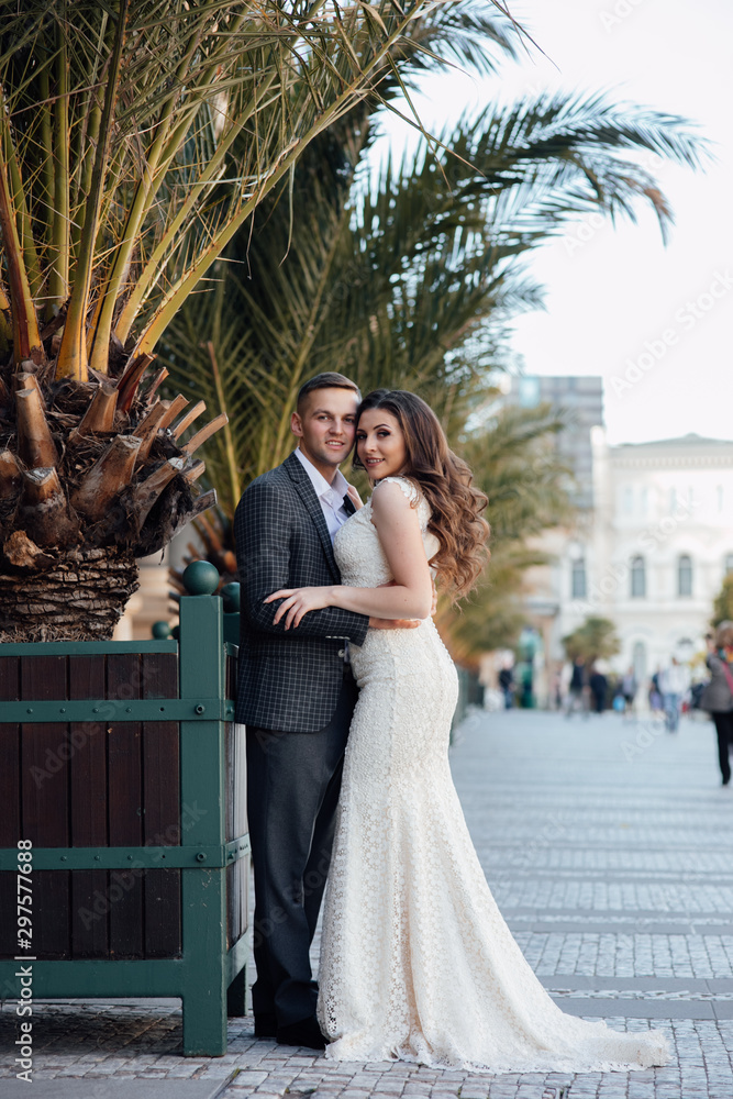 Naklejka premium Beautiful couple posing on streets near palm trees. Young sexy couple hugs near palm trees, enjoy their romantic vacation together. Couple wearing suit and white luxury dress, enjoy their honeymoon
