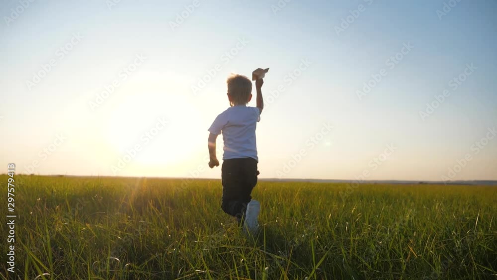 A small blond boy runs across the green, fresh grass and holds a paper airplane in his hands. The kid dreams of flying, he is very happy. A small blond boy runs across the green, fresh grass and holds a paper airplane in his hands. The kid dreams of flying, he is very happy.