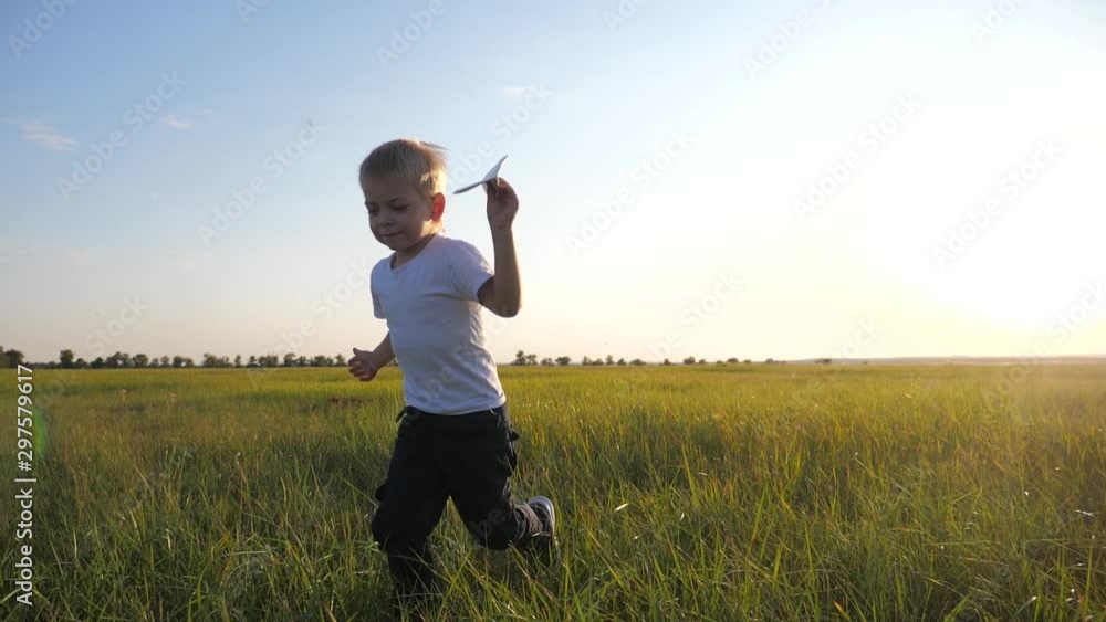 A small blond boy runs across the green, fresh grass and holds a paper airplane in his hands. The kid dreams of flying, he is very happy. A small blond boy runs across the green, fresh grass and holds a paper airplane in his hands. The kid dreams of flying, he is very happy.