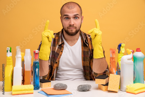 Isolated shot of bearded man has uncertain expression, stares at you, points to the top, sitting at the table with detergents isolated over yellow background photo